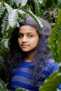 A young girl with curly hair stands amid vibrant green leaves, expressing calmness and natural beauty.